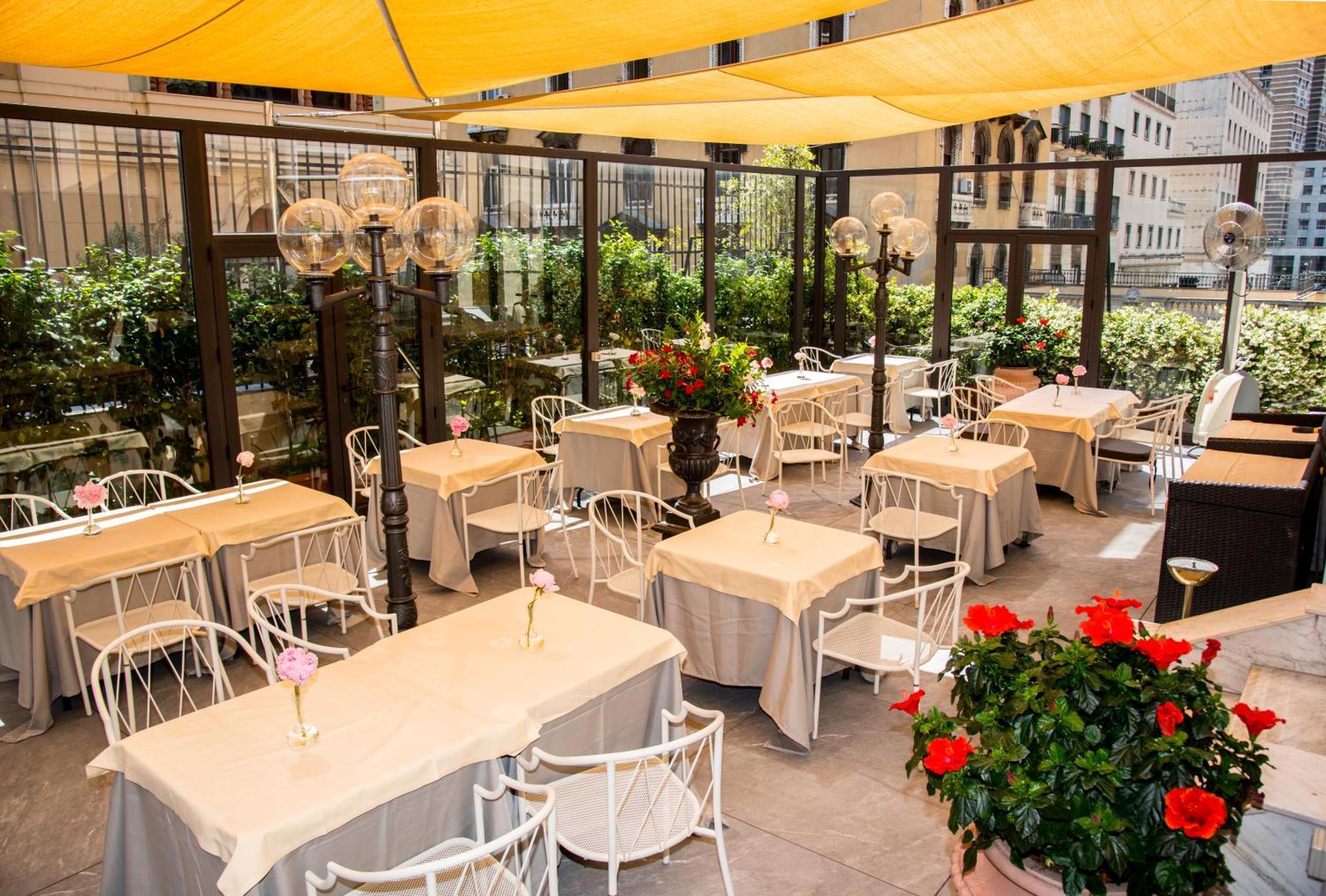 Hotel Bristol Palace Genova Esterno foto The photo shows an outdoor dining area, featuring several tables covered with light-colored tablecloths. Each table has a flower vase placed on it, adding a touch of decoration. The seating consists of white chairs, and the area is enclosed with glas