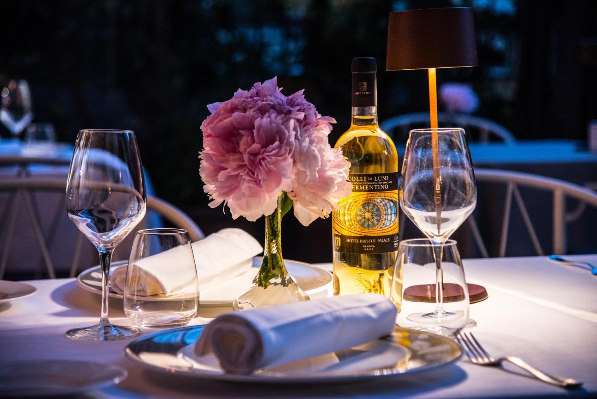 Hotel Bristol Palace Genova Esterno foto The photo shows a beautifully set dining table with an elegant ambiance. There is a vase with a pink flower arrangement at the center, adding a touch of color and freshness. A bottle of white wine is also prominently displayed next to the flowers. Th