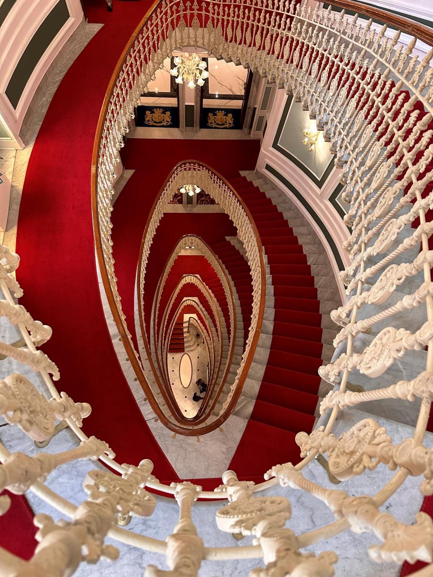Hotel Bristol Palace Genova Esterno foto The photo depicts a spiraling staircase viewed from above. The staircase features ornate, white railings and a striking red carpet that runs down the steps. The spiral design creates a visual effect of depth, leading the eye downwards. The surroundin