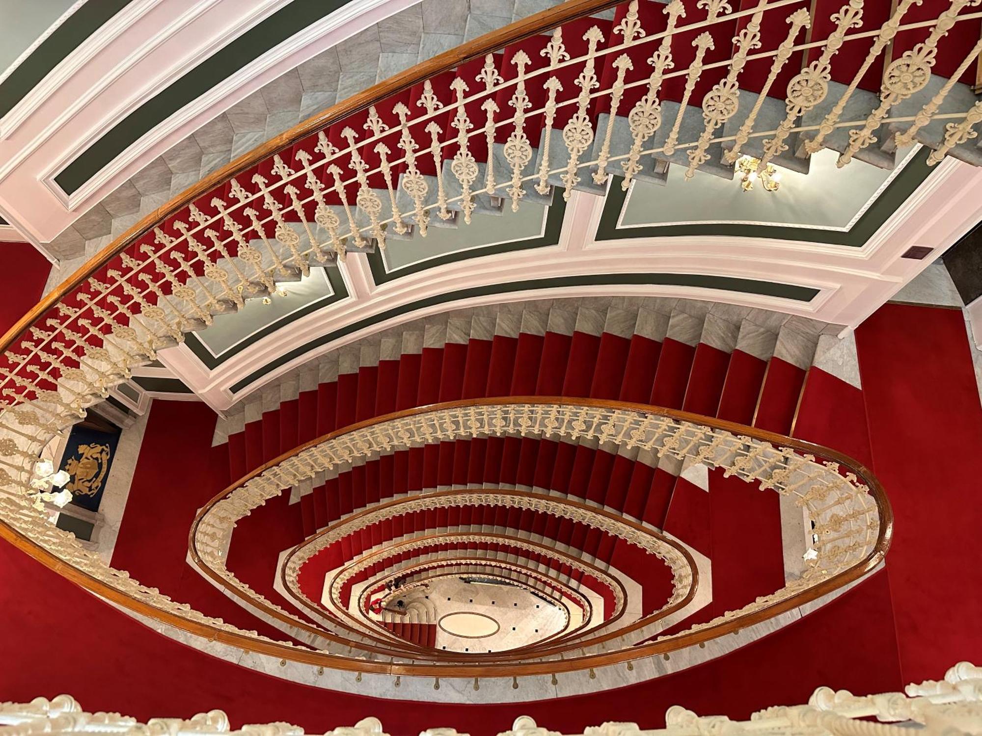 Hotel Bristol Palace Genova Esterno foto The photo shows a grand, spiral staircase viewed from above. The staircase features elegant white railings with intricate designs and is surrounded by a rich red carpet. The walls have a classic architectural style, with decorative elements in soft c