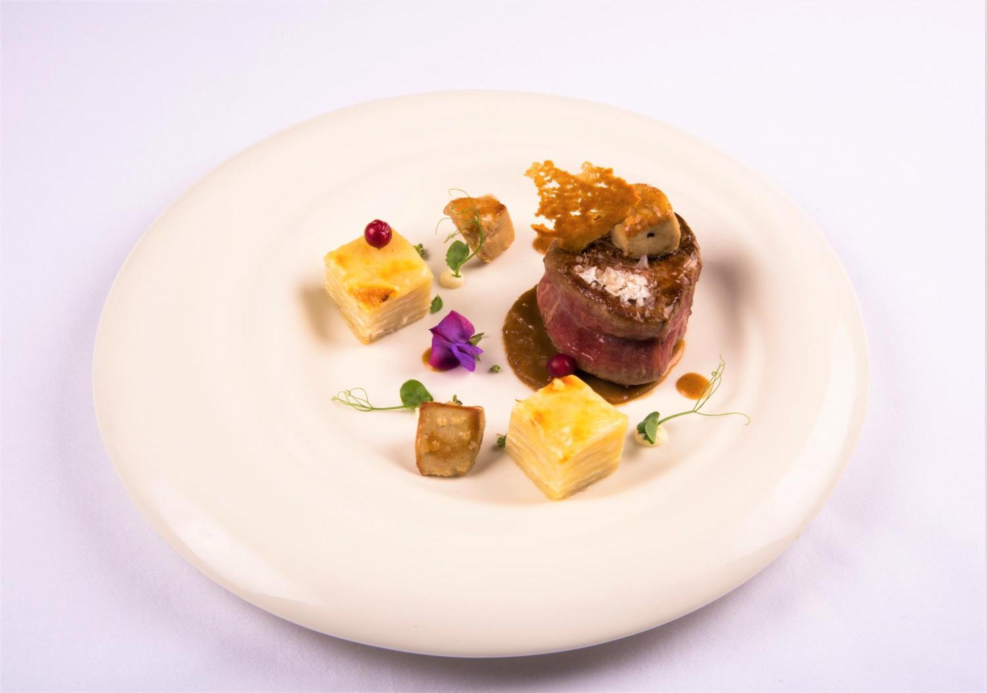 Hotel Bristol Palace Genova Esterno foto The photo shows an elegantly plated dish featuring a central piece of meat wrapped in a savory outer layer, possibly prosciutto or bacon, topped with a sauce or garnishment. Surrounding the main protein are small cubes that appear to be a starch, lik