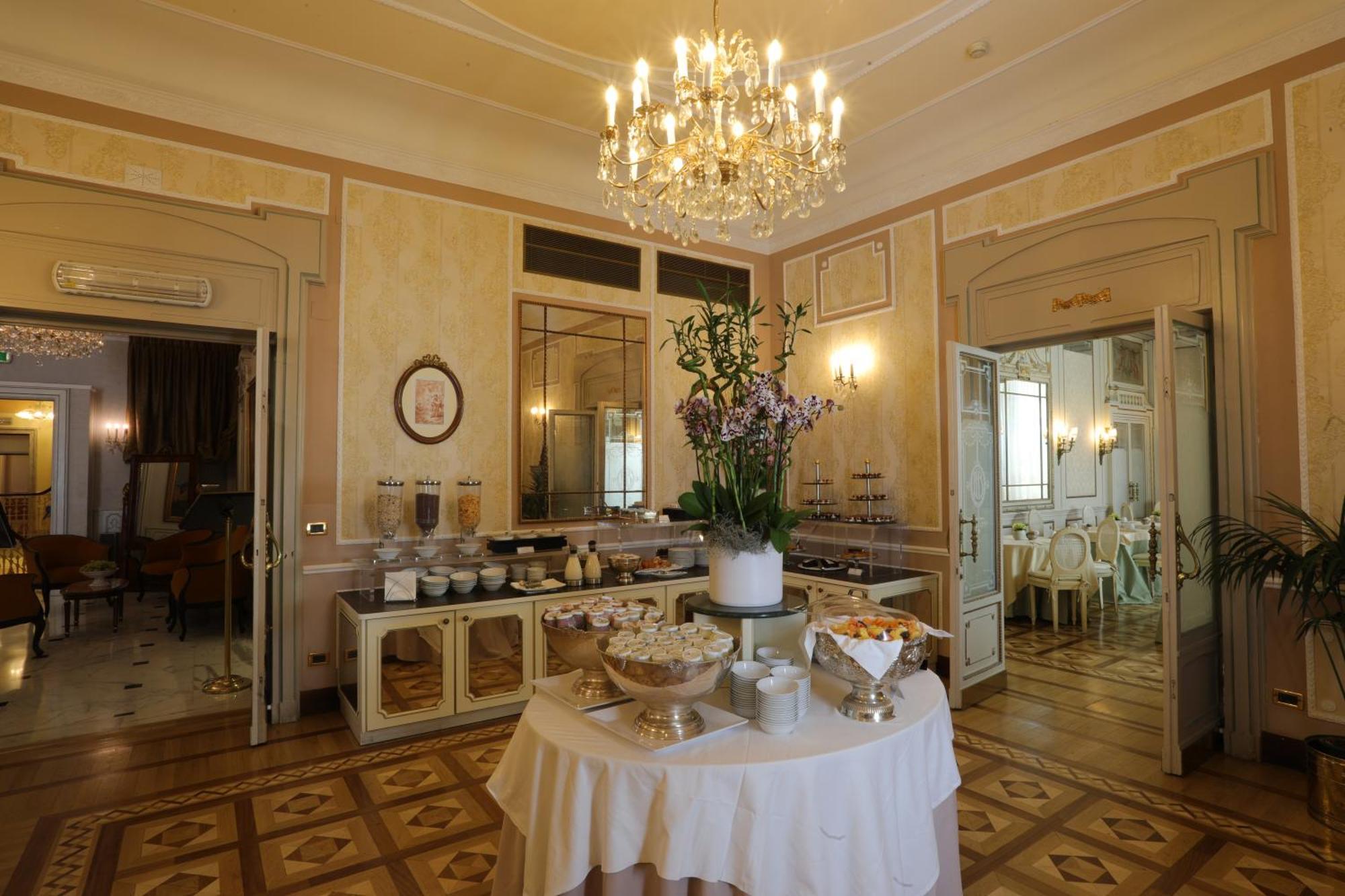 Hotel Bristol Palace Genova Esterno foto The photo shows an elegant interior of a dining area or event space. There is a beautifully decorated chandelier hanging from the ceiling, illuminating the room. In the center, there's a table draped with a white tablecloth, featuring a display of va