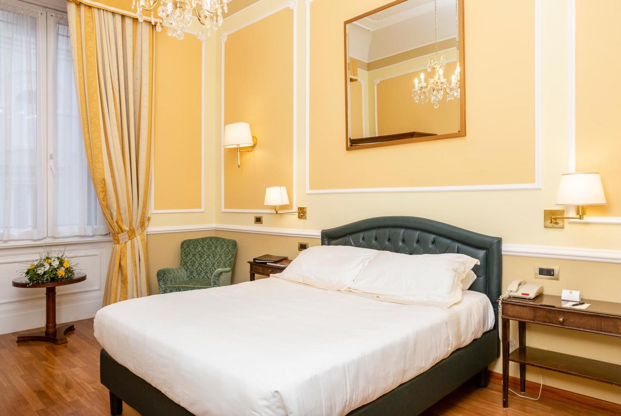 Hotel Bristol Palace Genova Camera foto The photo shows a cozy hotel room with a warm color scheme. It features a large bed with a white duvet, neatly arranged. To the left, there's a light green armchair and a small table. The walls are painted a soft yellow, and there are elegant curtain