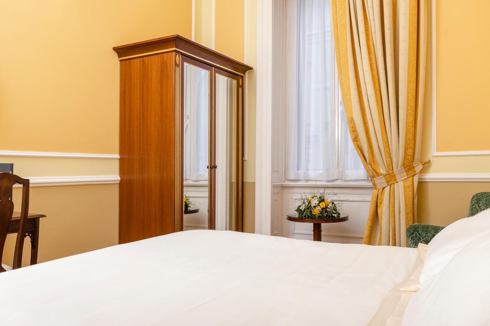 Hotel Bristol Palace Genova Camera foto The photo shows a cozy bedroom with warm yellow walls. There is a bed with white bedding in the foreground. On one side of the room, there is a wooden wardrobe that features a mirrored door. Beside it, there's a small table adorned with a floral arra