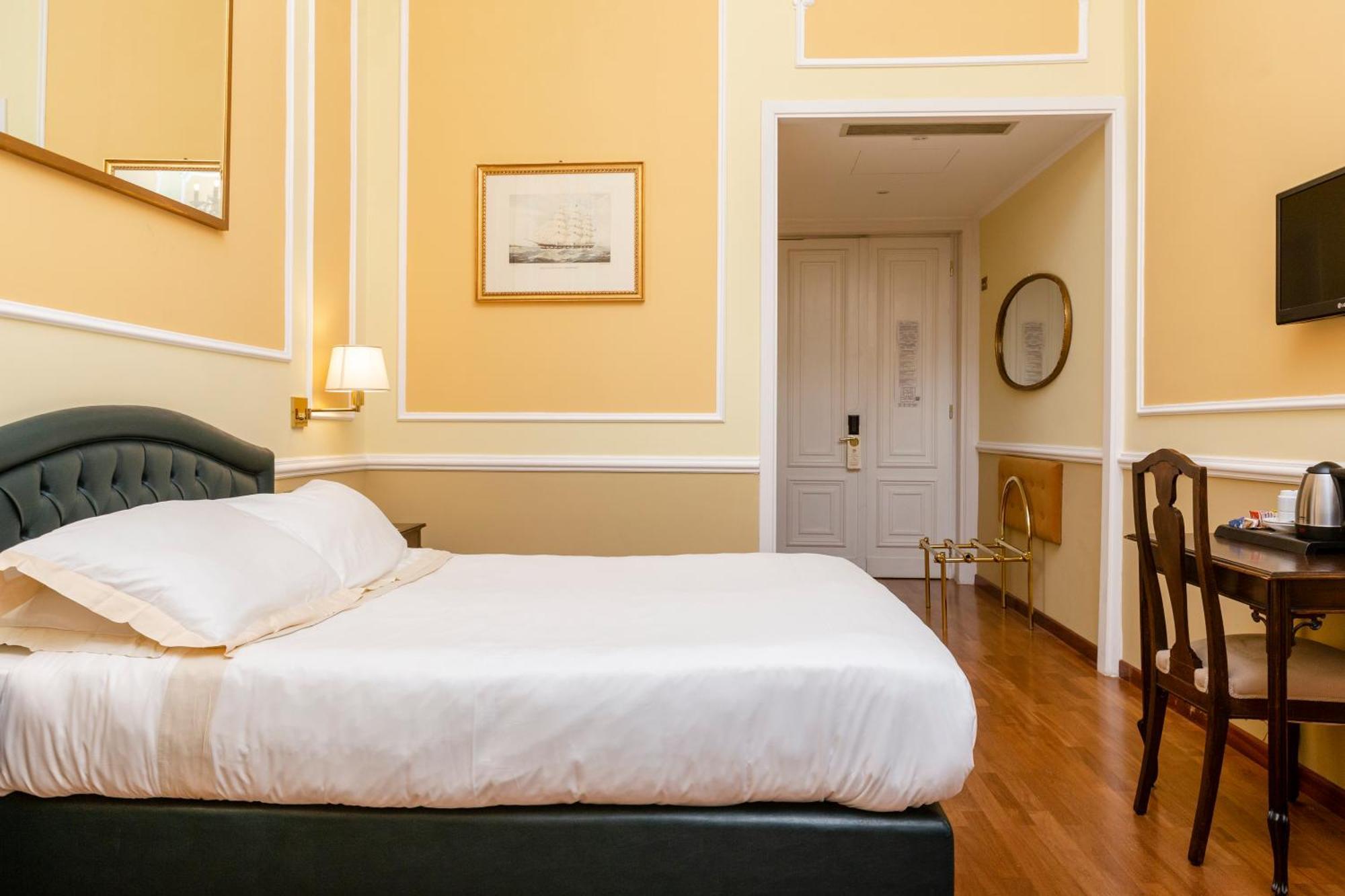 Hotel Bristol Palace Genova Camera foto The photo shows a cozy hotel room with a predominantly yellow interior. It features a large bed with white bedding and a tufted green headboard. Beside the bed, there are wall sconces providing soft lighting. On the walls, there are framed artworks, 