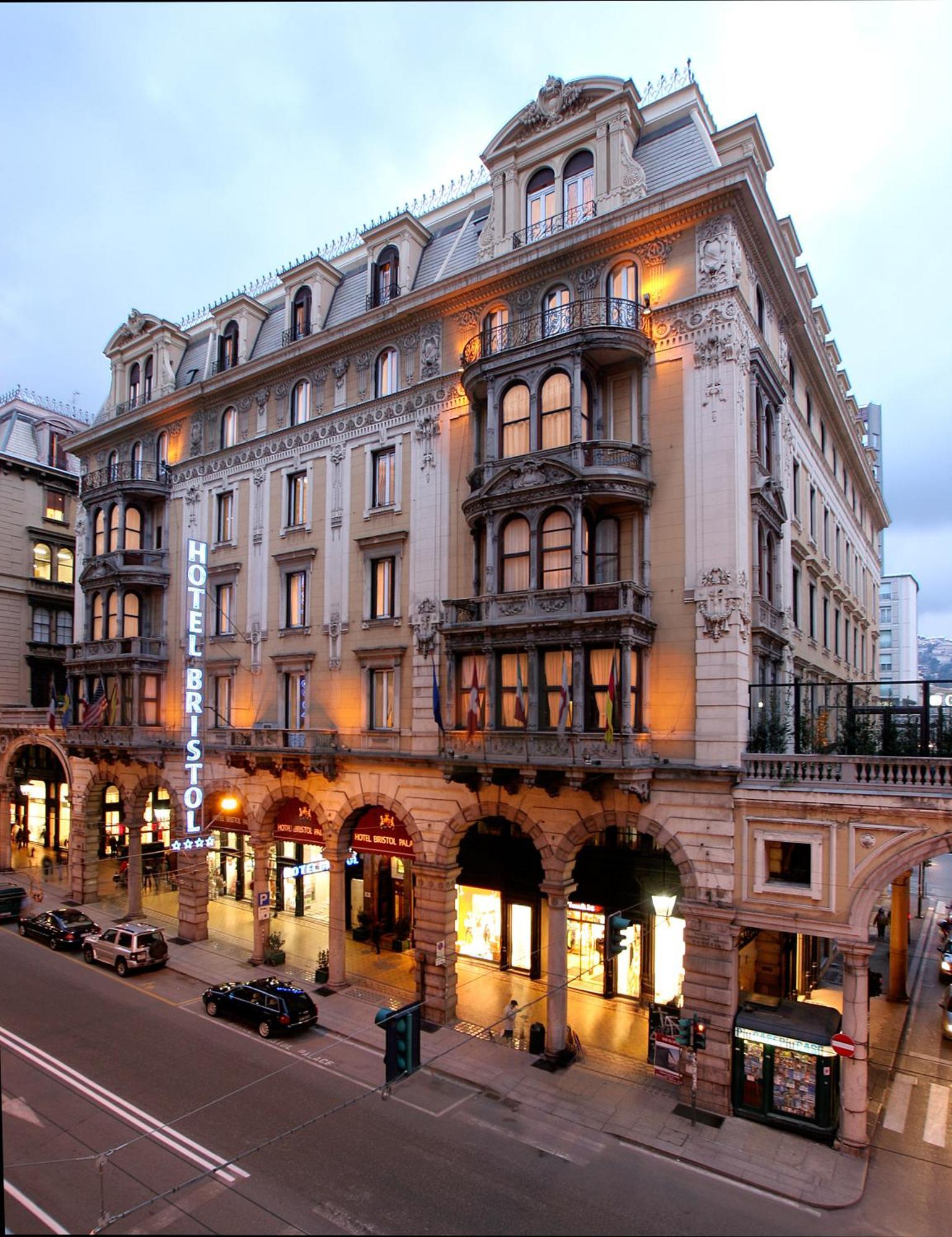 Hotel Bristol Palace Genova Esterno foto The photo shows a grand hotel building, likely the Hotel Bristol. It features an elegant architectural design with ornate details, including balconies and decorative moldings. The façade is illuminated, giving it a warm glow in the evening light. The