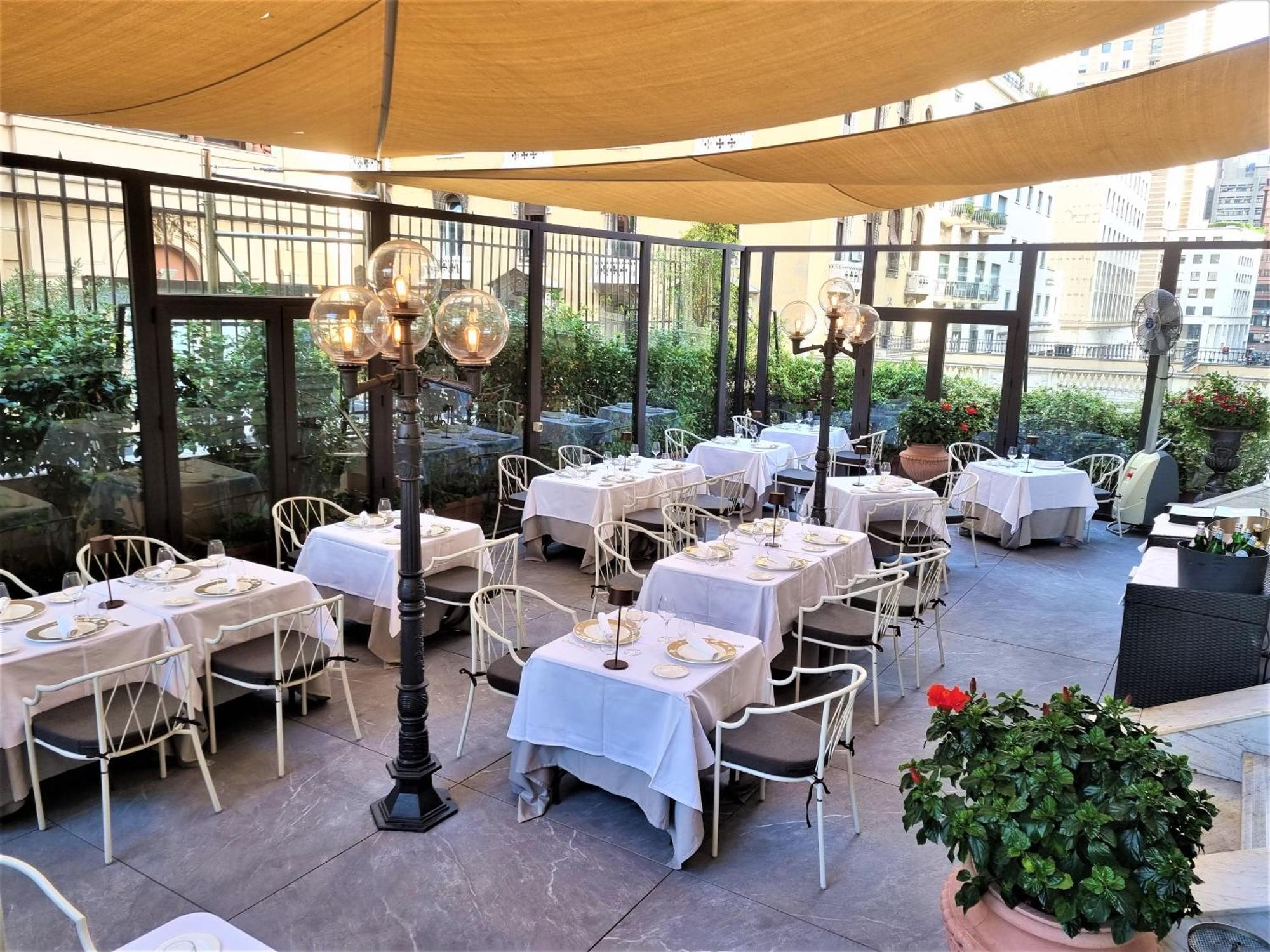Hotel Bristol Palace Genova Esterno foto The photo shows an outdoor dining area of a restaurant. There are several neatly arranged tables covered with white tablecloths, set with plates and utensils. The space is adorned with decorative lanterns on tall stands, providing a cozy atmosphere. 