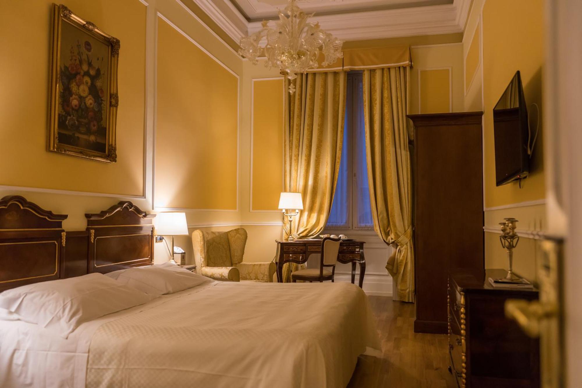 Hotel Bristol Palace Genova Camera foto The photo shows a beautifully designed bedroom with warm yellow walls. There is a large bed with a white bedspread positioned against one wall. Next to the bed, there are two table lamps providing soft lighting. A cozy seating area is visible, featur
