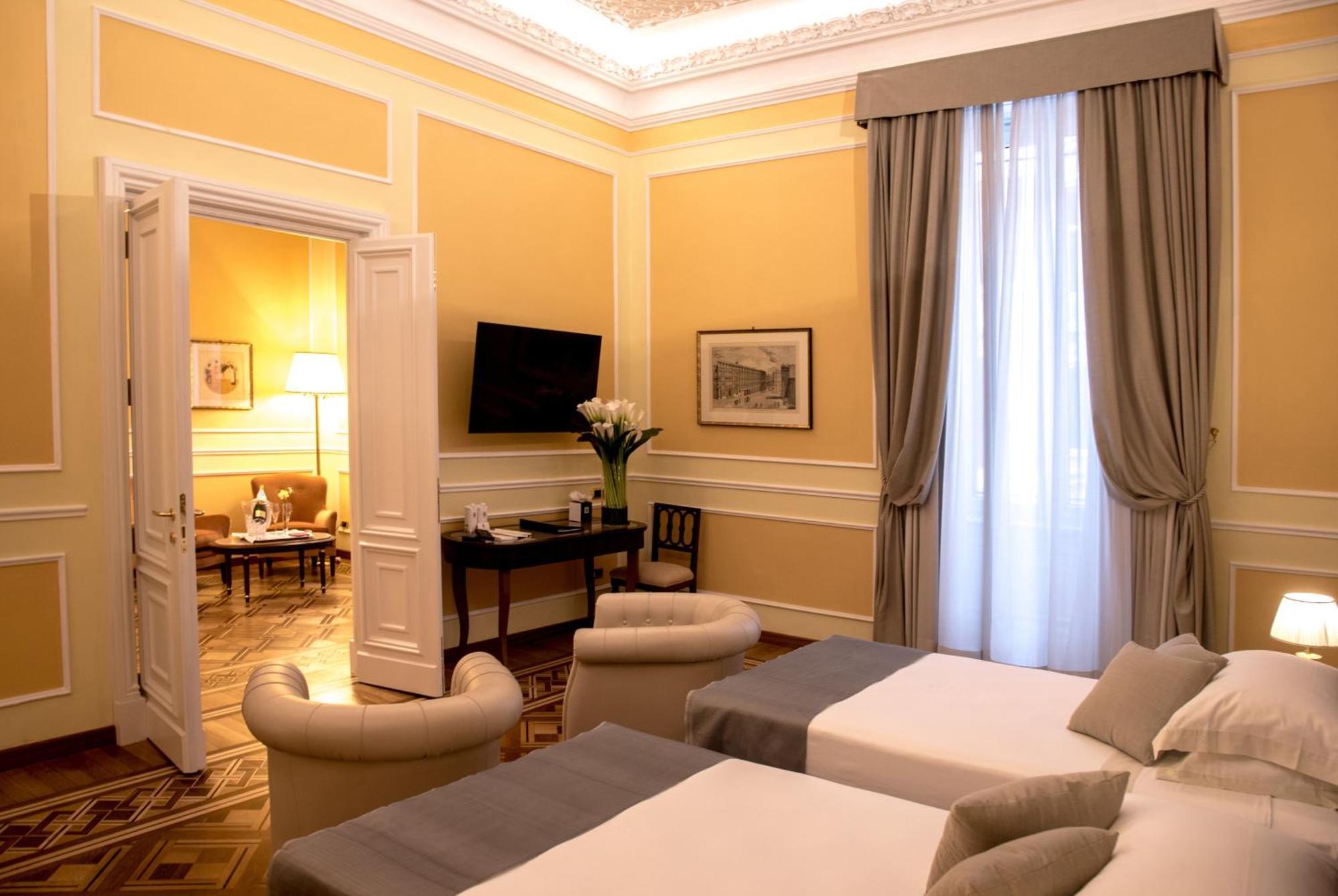 Hotel Bristol Palace Genova Esterno foto The photo shows an elegantly designed hotel room with soft yellow walls and high ceilings. There are two beds with gray covers positioned in the foreground. The room features large windows with flowing curtains that allow natural light to enter. In t