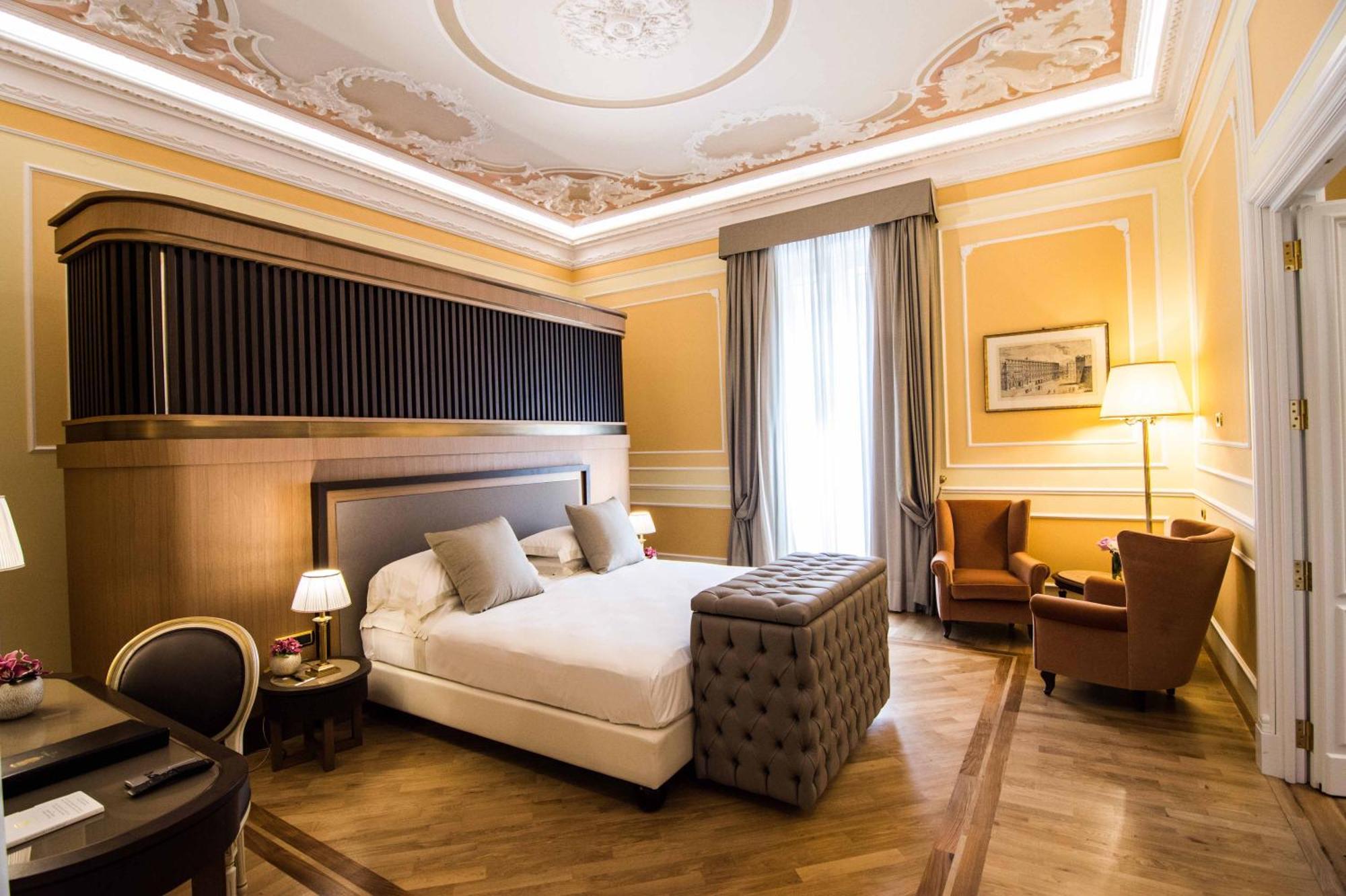 Hotel Bristol Palace Genova Esterno foto The photo shows an elegantly designed hotel room. It features a large bed with plush pillows, and a tufted ottoman at the foot of the bed. The walls are painted in a warm yellow tone, adorned with decorative molding. A pair of tall windows are draped
