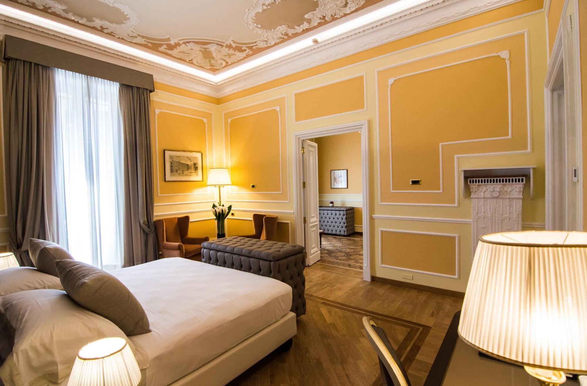 Hotel Bristol Palace Genova Esterno foto The photo shows a well-decorated hotel room with a warm yellow color scheme. There is a large bed with neutral-toned bedding and pillows. To the side, there are two armchairs and a small table, creating a cozy seating area. A stylish lamp sits on a n