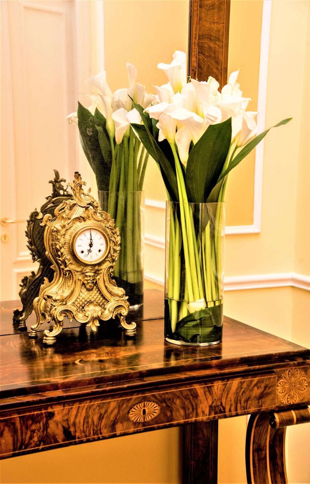 Hotel Bristol Palace Genova Esterno foto The photo features an ornate vintage clock with a decorative gold finish, placed on a wooden surface. Next to the clock is a tall, clear vase filled with elegant white calla lilies and green leaves. The background shows a softly lit interior with lig