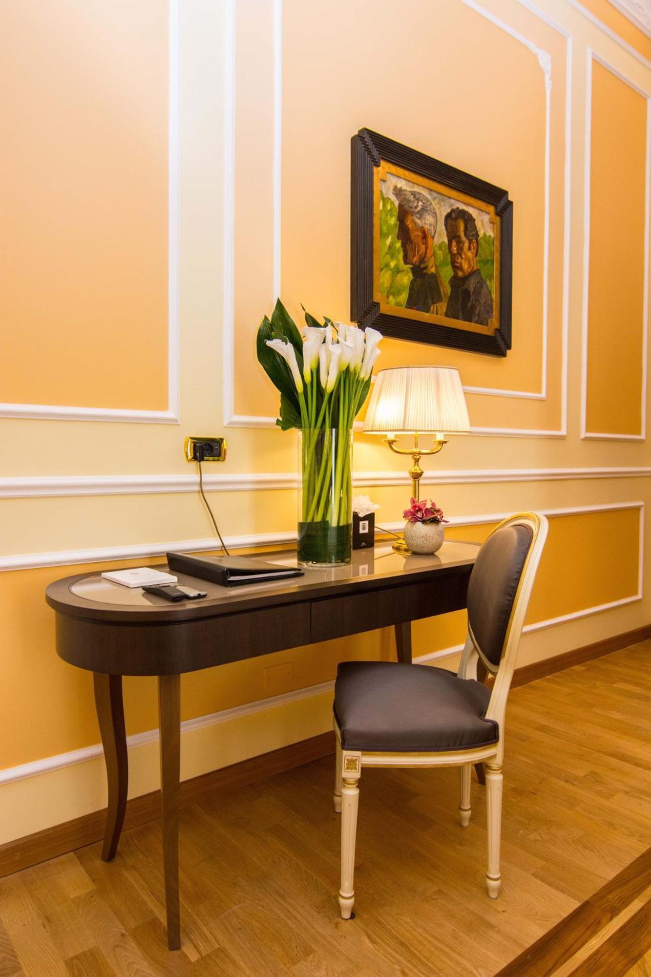 Hotel Bristol Palace Genova Esterno foto The photo shows a stylishly arranged corner of a room. There is a sleek, curved desk made of dark wood positioned against a softly colored wall. On the desk, there is a black folder and a small decorative item, possibly a flower pot. A tall, elegant 