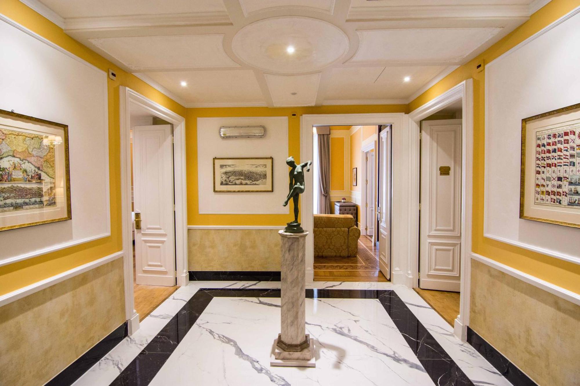 Hotel Bristol Palace Genova Esterno foto The photo shows an elegant hallway with a bright yellow wall and marble flooring. The ceiling is detailed, featuring a circular design. In the center, there is a statue on a small pedestal, adding a decorative touch to the space. The walls are adorne