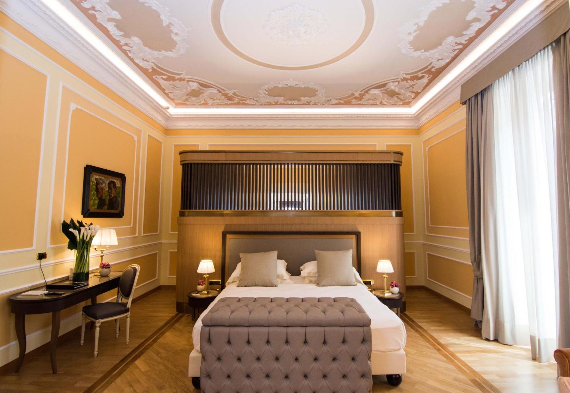 Hotel Bristol Palace Genova Esterno foto The photo shows a stylish and elegant bedroom. The room features light peach-colored walls with ornate moldings. The ceiling has decorative elements, creating an upscale atmosphere. In the center, there is a large, gray upholstered bed with plush bed