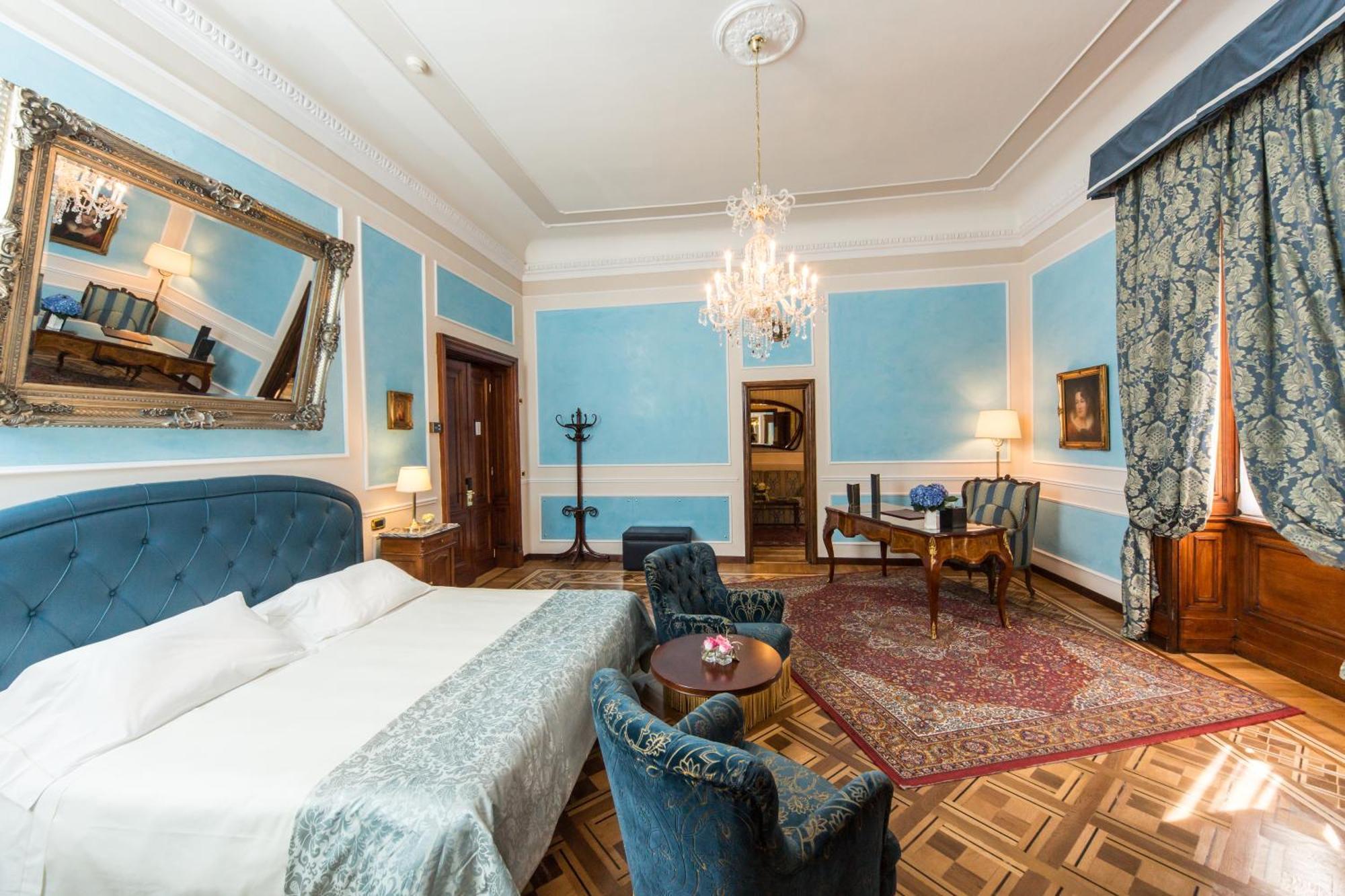 Hotel Bristol Palace Genova Camera foto The photo shows a beautifully designed hotel room or suite. It features light blue walls with decorative molding, providing an elegant ambiance. The room includes a large bed with a gray coverlet, accompanied by plush blue armchairs and a round table