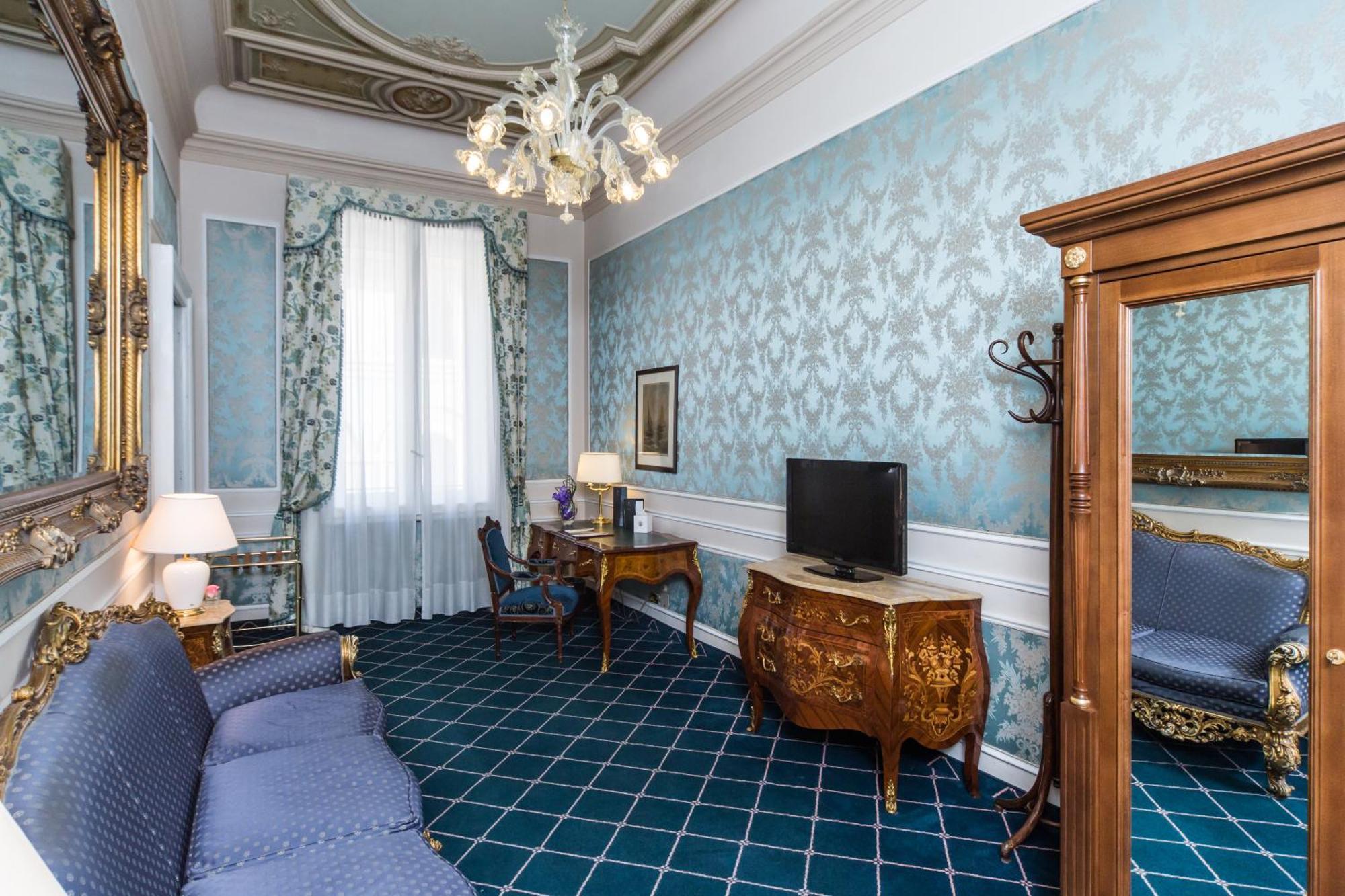 Hotel Bristol Palace Genova Camera foto The photo shows an elegantly furnished room with a light blue and patterned wall. There is a decorative chandelier hanging from the ceiling. On the left, there is a blue sofa, and next to it is a lamp providing additional lighting. A wooden desk with