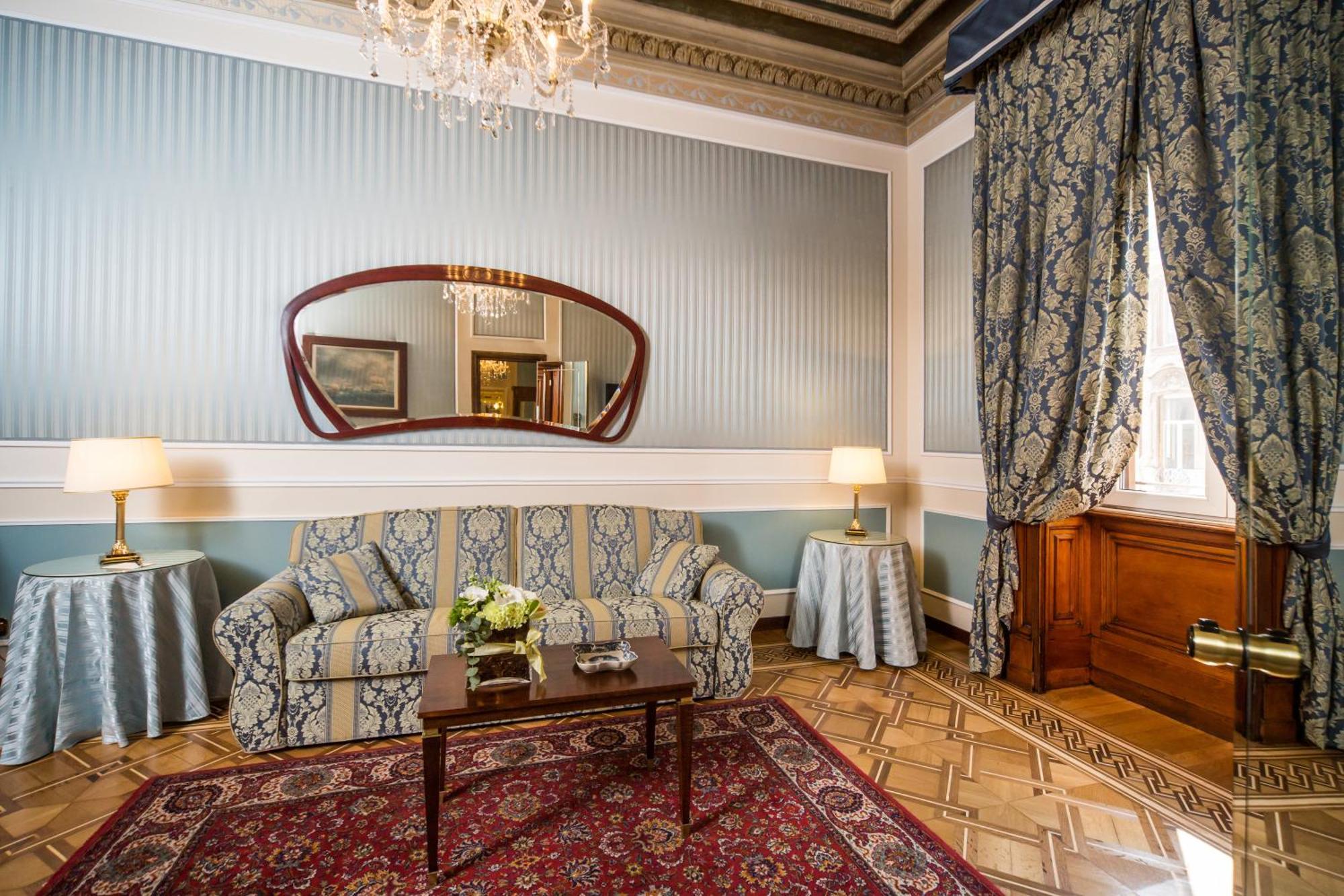 Hotel Bristol Palace Genova Camera foto The photo shows an elegant living room featuring classic decor. The walls are painted in a soft blue with vertical stripes, and ornate crown molding adds to the room's charm. A large mirror with a wavy frame hangs above a patterned couch that complem