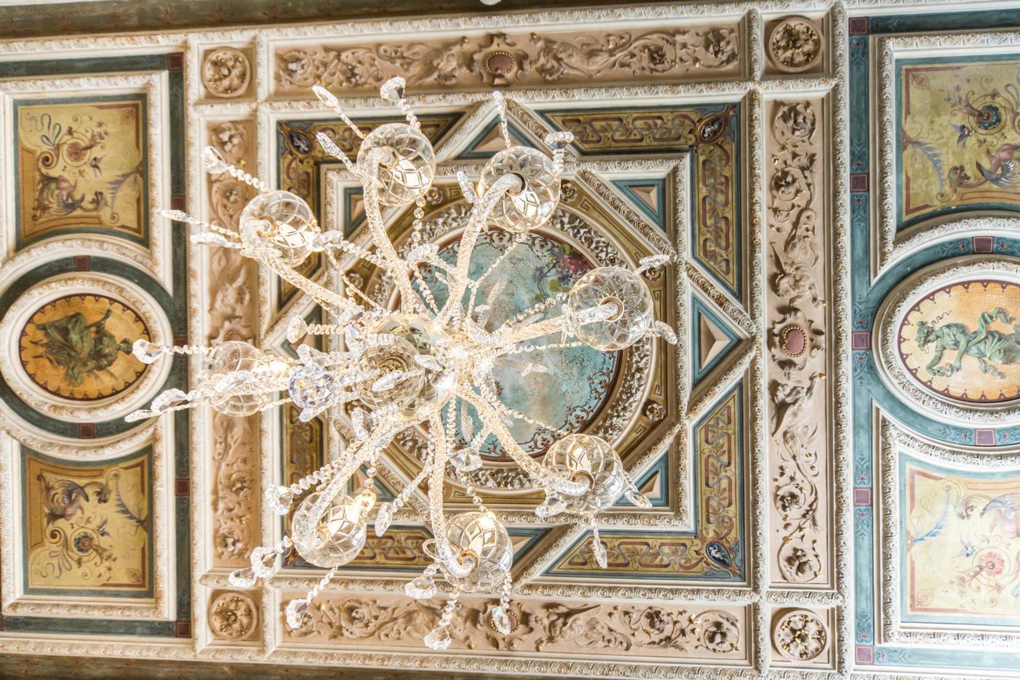 Hotel Bristol Palace Genova Camera foto The image depicts an ornate ceiling featuring a large chandelier made of crystal. The chandelier hangs from a richly detailed ceiling adorned with intricate designs, including decorative molding and colorful frescoes. The artwork includes circular mo